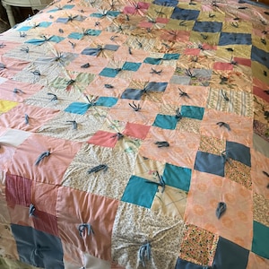 Puede incluir: Un edredón de retazos con una variedad de telas en patrones rosas, azules y florales. El edredón está hecho de parches cuadrados atados con hilo azul. El edredón está sobre una cama con un cabecero de metal blanco.
