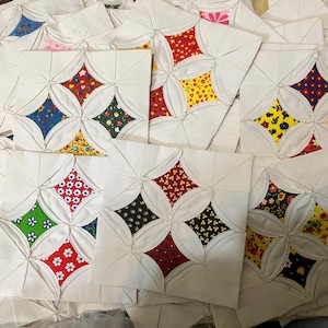 May include: A collection of white fabric squares with a four-petal design, each petal filled with colorful floral and patterned fabrics. The squares are arranged in a pile, showcasing various color combinations.