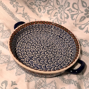 Puede incluir: Fuente redonda de cerámica para hornear con un estampado floral azul y blanco. El plato tiene un borde y asas de color azul oscuro. El interior presenta un diseño de hojas azules repetidas sobre un fondo blanco. El borde está decorado con un patrón marrón y verde.