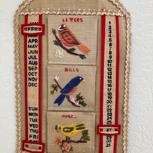 May include: Vintage wall organizer made of woven material with embroidered bird designs. Features pockets labeled "Letters", "Bills", and "Misc.", along with a calendar grid and month/day indicators. The organizer is trimmed with a woven border and red accents.