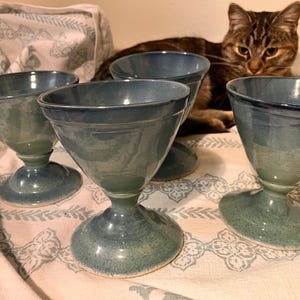 Pode incluir: Quatro taças de sobremesa de cerâmica azul-esverdeada com tigelas alargadas e bases de pedestal são dispostas sobre um tecido estampado. As taças têm um acabamento brilhante e um gradiente de cor. Um gato malhado está no fundo.
