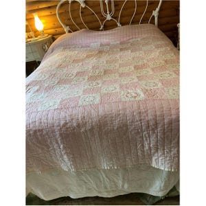 Könnte beinhalten: Eine rosa-weiße Steppdecke mit floralem Muster bedeckt ein Bett. Die Steppdecke hat ein Rasterdesign mit abwechselnden rosa und weißen Quadraten und einen weißen Spitzen-Bettrock. Das Bett hat ein weißes Metall-Kopfteil.