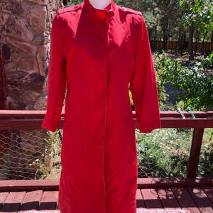 May include: A bright red, long coat with a stand-up collar and button details on the shoulders. The coat is displayed on a mannequin, showcasing its full length and design. The coat is a solid color and appears to be made of a smooth fabric.