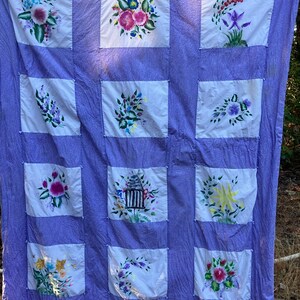 May include: A lavender quilt featuring twelve white square panels, each decorated with hand-painted floral arrangements and a central birdcage illustration. The fabric has a subtle textured pattern and is joined by small fabric ties at each corner.