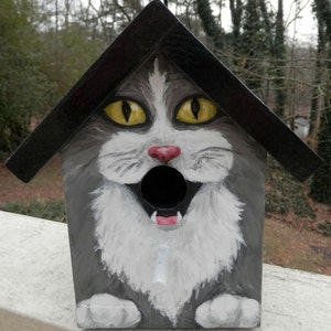 May include: A gray and white cat face painted on a black wooden birdhouse. The cat has yellow eyes, a red nose, and an open mouth with white teeth. The birdhouse has a black roof and a round hole for birds to enter.