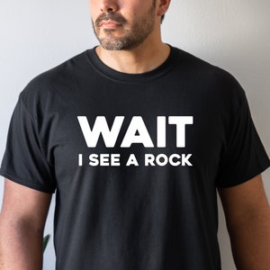 May include: Black t-shirt with the white text "WAIT I SEE A ROCK". The shirt is a crew neck style and appears to be made of a soft material. The text is centered on the chest.