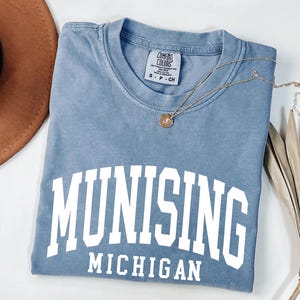 May include: A blue short-sleeved t-shirt with a crew neckline and a white screen-printed graphic that reads MUNISING MICHIGAN in a bold, arched collegiate font. The garment features a Comfort Colors brand label at the inner neck collar.