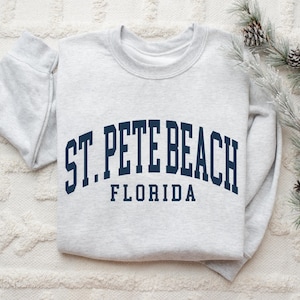 May include: A light gray sweatshirt with the text "ST. PETE BEACH FLORIDA" in navy blue arched lettering. The sweatshirt is folded and rests on a white textured surface, with pine branches and cones in the background.