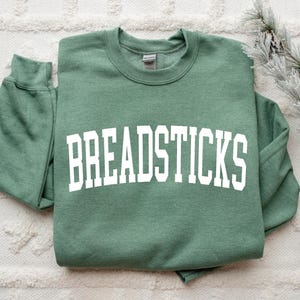 May include: A sage green sweatshirt with the word "BREADSTICKS" printed in large, white, block letters. The sweatshirt is folded neatly on a white, textured surface. A sprig of faux pine with pinecones is in the background.