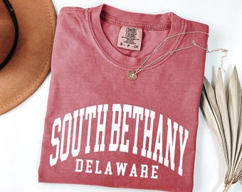 Puede incluir: Una camiseta de manga corta color rosa polvoriento con el texto blanco SOUTH BETHANY DELAWARE en una fuente universitaria. La camiseta tiene cuello redondo y una etiqueta de la marca Comfort Colors en el interior. Una fina cadena dorada descansa sobre la tela.
