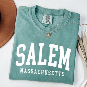 Puede incluir: Una camiseta verde agua con la palabra "SALEM" en letras blancas grandes y "MASSACHUSETTS" debajo. Un collar dorado con un colgante y un sombrero marr&oacute;n est&aacute;n al lado de la camiseta. La camiseta est&aacute; doblada cuidadosamente.