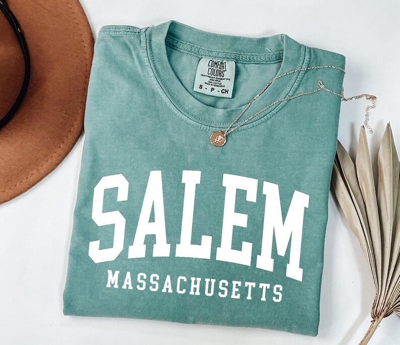 Puede incluir: Una camiseta verde agua con la palabra "SALEM" en letras blancas grandes y "MASSACHUSETTS" debajo. Un collar dorado con un colgante y un sombrero marr&oacute;n est&aacute;n al lado de la camiseta. La camiseta est&aacute; doblada cuidadosamente.
