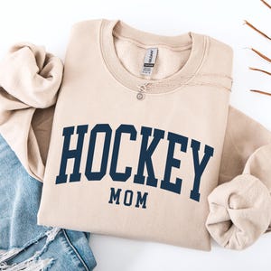 Op de afbeelding: Een beige sweatshirt met de tekst "HOCKEY MOM" in marineblauw. De sweatshirt is opgevouwen, met de mouwen naar binnen gevouwen. Een zilveren ketting met een kleine hanger ligt op de sweatshirt. Een blauwe jeans is zichtbaar op de achtergrond.