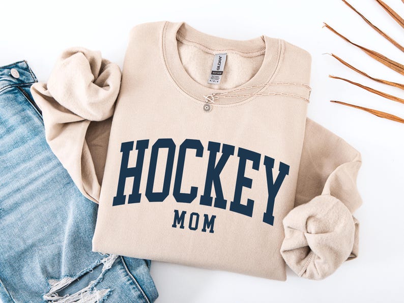 Op de afbeelding: Een beige sweatshirt met de tekst "HOCKEY MOM" in marineblauw. De sweatshirt is opgevouwen, met de mouwen naar binnen gevouwen. Een zilveren ketting met een kleine hanger ligt op de sweatshirt. Een blauwe jeans is zichtbaar op de achtergrond.