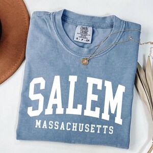 Puede incluir: Una camiseta azul con la palabra "SALEM" en letras blancas grandes, con "MASSACHUSETTS" debajo. Un collar dorado con un colgante descansa sobre la camiseta. Un sombrero marr&oacute;n y una planta seca est&aacute;n en el fondo.