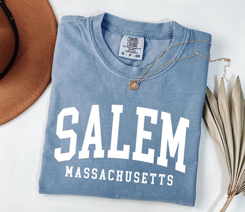 Puede incluir: Una camiseta azul con la palabra "SALEM" en letras blancas grandes, con "MASSACHUSETTS" debajo. Un collar dorado con un colgante descansa sobre la camiseta. Un sombrero marr&oacute;n y una planta seca est&aacute;n en el fondo.
