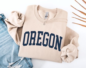 May include: A beige sweatshirt with the word "OREGON" printed in navy blue block letters. The sweatshirt is folded, with a pair of blue jeans visible on the left. A silver necklace with a small pendant is on the sweatshirt.