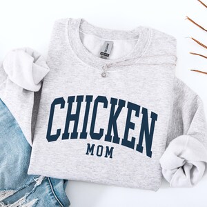 May include: A light gray sweatshirt with the words "CHICKEN MOM" in navy blue block letters. The sweatshirt is folded with the sleeves rolled up. A silver necklace with a small pendant is on the sweatshirt. Blue jeans are visible in the background.