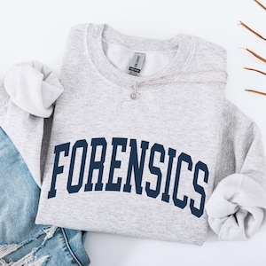 May include: A light gray sweatshirt with the word "FORENSICS" in navy blue block letters across the chest. The sweatshirt is folded, with the sleeves and a pair of blue jeans visible. A silver necklace with a small pendant is on the sweatshirt.