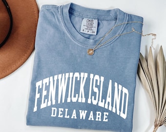 Puede incluir: Una camiseta azul de manga corta con cuello redondo que presenta el texto blanco FENWICK ISLAND DELAWARE en una fuente de estilo universitario. La camiseta tiene una etiqueta de la marca Comfort Colors en el cuello y es talla S.