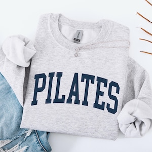 May include: A light gray Pilates sweatshirt with navy blue lettering, paired with blue jeans. A silver necklace with a small pendant rests on the sweatshirt. The sleeves are cuffed. Dried palm leaves are in the background.