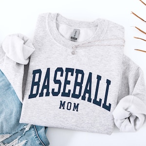 May include: A light gray sweatshirt with the words "BASEBALL MOM" in navy blue arched across the front. The sweatshirt is folded with the sleeves rolled up. A silver necklace and blue jeans are also in the image.