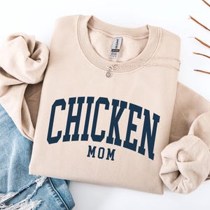 May include: A beige sweatshirt with the words "CHICKEN MOM" in navy blue arched lettering. The sweatshirt is folded with a pair of blue jeans and a necklace. Dried leaves are in the background.
