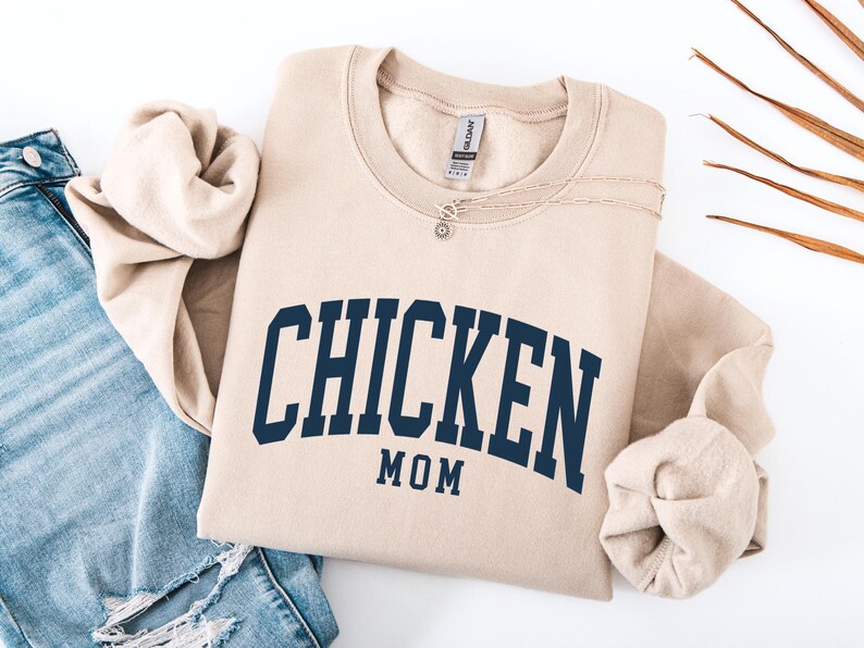 May include: A beige sweatshirt with the words "CHICKEN MOM" in navy blue arched lettering. The sweatshirt is folded with a pair of blue jeans and a necklace. Dried leaves are in the background.