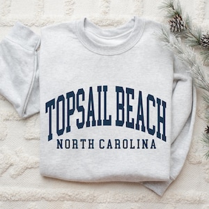 May include: A pale grey sweatshirt featuring the navy blue text "TOPSAIL BEACH NORTH CAROLINA" in an arched design. The garment is folded on a white textured surface, with pine branches and cones in the background.