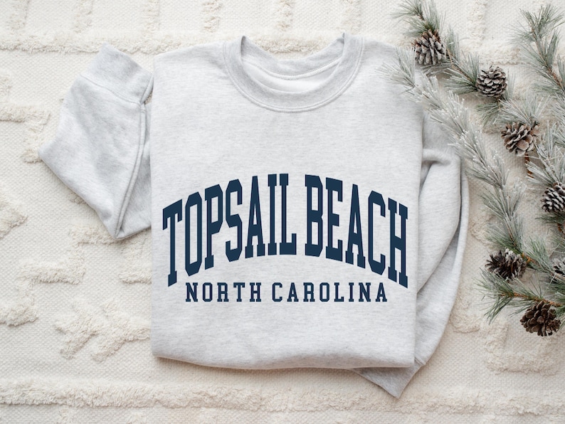 May include: A pale grey sweatshirt featuring the navy blue text "TOPSAIL BEACH NORTH CAROLINA" in an arched design. The garment is folded on a white textured surface, with pine branches and cones in the background.