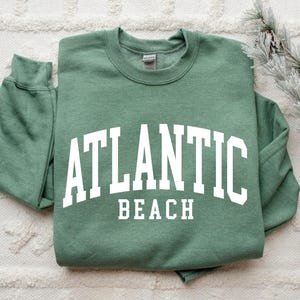 May include: Sage green crewneck sweatshirt with the words "ATLANTIC BEACH" in bold white lettering. The sweatshirt has long sleeves and a classic, casual style. Perfect for a beach vacation or everyday wear.