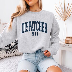 May include: A light gray crewneck sweatshirt featuring the dark navy blue text DISPATCHER 911 printed in a collegiate-style arched font across the chest. The garment has long sleeves and a ribbed neckline, cuffs, and hem, providing a classic fit.