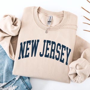 May include: A beige sweatshirt with the text "NEW JERSEY" in navy blue, arched across the chest. The sweatshirt is folded, with a silver necklace and a pair of blue jeans visible.