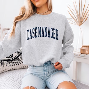 May include: A light gray crewneck sweatshirt featuring the text CASE MANAGER printed in a dark navy blue arched font across the chest. The garment has long sleeves and a ribbed collar, cuffs, and hem, providing a classic and comfortable fit.