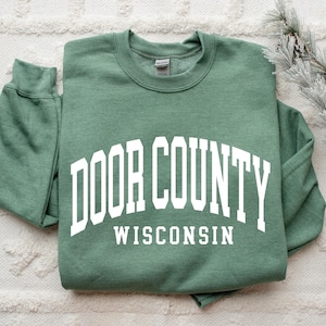 May include: A sage green crewneck sweatshirt with the words "DOOR COUNTY WISCONSIN" in white block letters. The sweatshirt is folded and lies on a white textured surface. A sprig of faux pine and pinecones is in the background.