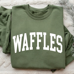 May include: Olive green sweatshirt with the word "WAFFLES" in large, white, arched letters across the front. The sweatshirt is folded, with the sleeves visible. The background includes a white, textured surface and decorative pine branches.