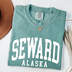 May include: A sage green short-sleeved t-shirt with a crew neckline. The shirt features the white text SEWARD ALASKA printed in a bold, arched block font across the chest. A Comfort Colors brand label is visible at the inner neck collar.