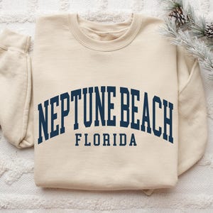 May include: Cream-colored sweatshirt with the text "NEPTUNE BEACH FLORIDA" in navy blue arched lettering. The sweatshirt has a crew neck and long sleeves. The image is taken from above, with a sprig of pine needles and pine cones in the upper right corner.