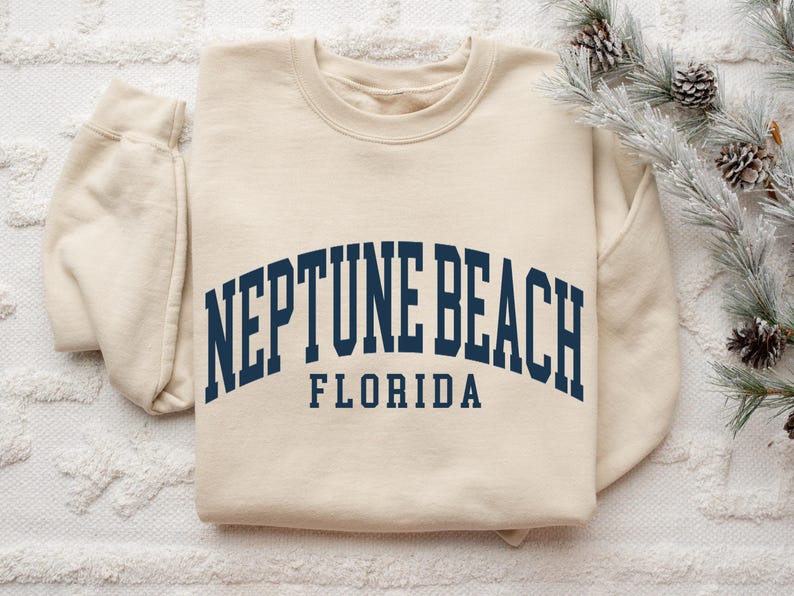 May include: Cream-colored sweatshirt with the text "NEPTUNE BEACH FLORIDA" in navy blue arched lettering. The sweatshirt has a crew neck and long sleeves. The image is taken from above, with a sprig of pine needles and pine cones in the upper right corner.