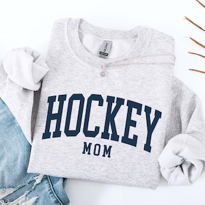 Op de afbeelding: Een lichtgrijs sweatshirt met de marineblauwe tekst "HOCKEY MOM" in een gebogen ontwerp. De mouwen zijn opgerold. Een zilveren ketting met een kleine hanger ligt op de trui. Een blauwe jeans is zichtbaar in de afbeelding.