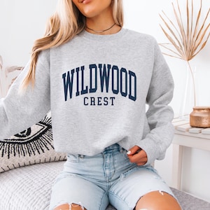 May include: A light gray sweatshirt with the text "WILDWOOD CREST" in navy blue block letters. The sweatshirt is a crew neck style and is made of a soft material. The sweatshirt is paired with light blue denim shorts.