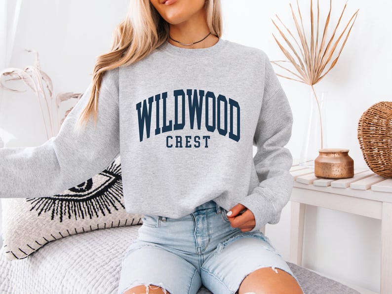 May include: A light gray sweatshirt with the text "WILDWOOD CREST" in navy blue block letters. The sweatshirt is a crew neck style and is made of a soft material. The sweatshirt is paired with light blue denim shorts.