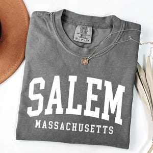 Puede incluir: Una camiseta gris con la palabra "SALEM" en letras blancas grandes, con "MASSACHUSETTS" debajo. Un collar dorado con un colgante descansa sobre la camiseta. Un sombrero marr&oacute;n y una planta seca est&aacute;n en el fondo.