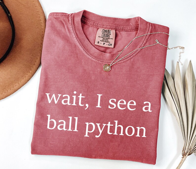 K&ouml;nnte beinhalten: Ein altrosa T-Shirt mit kurzen &Auml;rmeln und Rundhalsausschnitt. Auf der Brust befindet sich der wei&szlig;e Schriftzug "wait, I see a ball python". Das Etikett im Nackenbereich zeigt den Markennamen "Comfort Colors" und die Gr&ouml;&szlig;e S.