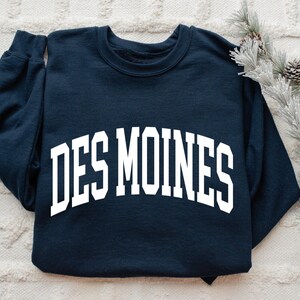May include: Navy blue sweatshirt with the white text "DES MOINES" in an arched design. The long-sleeved top is displayed on a white textured surface, with pinecones and greenery in the background.