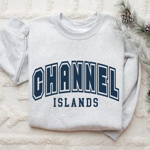 Pu&ograve; includere: Una felpa grigio chiaro con la scritta "CHANNEL ISLANDS" in lettere maiuscole blu navy. La felpa piegata &egrave; appoggiata su una superficie bianca strutturata, con un rametto di aghi di pino e pigne sullo sfondo.
