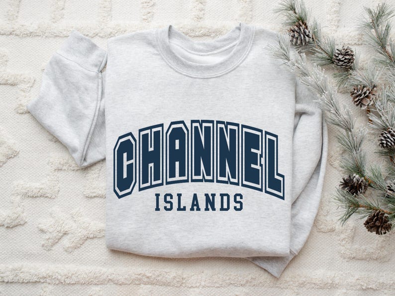 Pu&ograve; includere: Una felpa grigio chiaro con la scritta "CHANNEL ISLANDS" in lettere maiuscole blu navy. La felpa piegata &egrave; appoggiata su una superficie bianca strutturata, con un rametto di aghi di pino e pigne sullo sfondo.