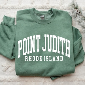 May include: A sage green sweatshirt with the words "POINT JUDITH RHODE ISLAND" printed in white block letters. The sweatshirt is folded neatly on a white textured surface. A sprig of faux pine and pinecones are in the background.