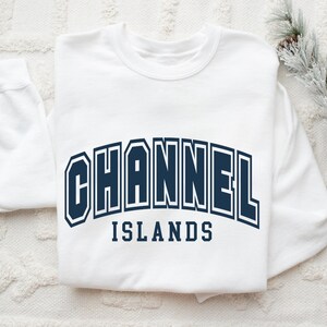 Pu&ograve; includere: Una felpa bianca con la scritta "CHANNEL ISLANDS" in lettere blu navy. Il testo &egrave; arcuato sul petto. La felpa &egrave; su una superficie bianca strutturata con pigne e vegetazione sullo sfondo.