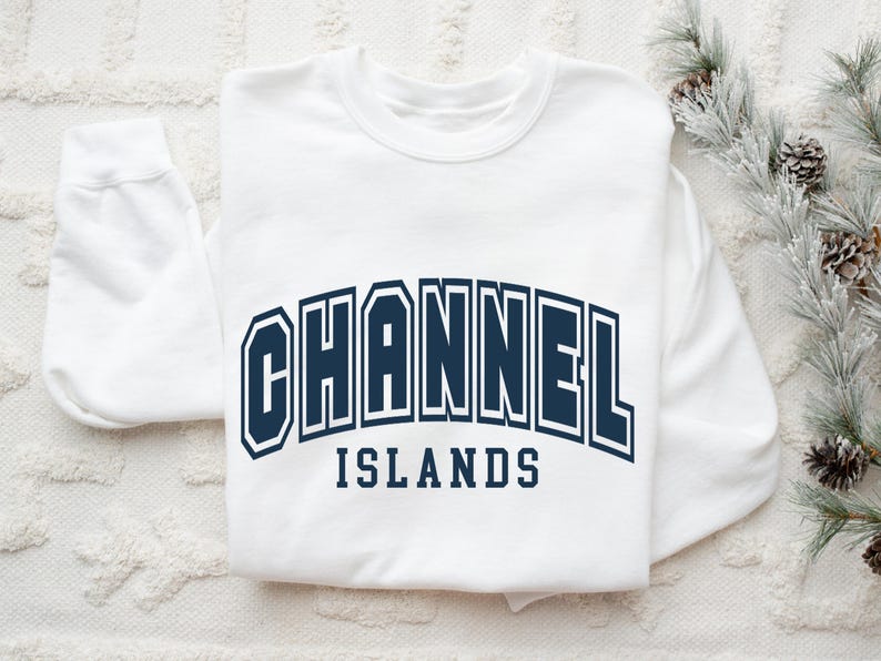 Pu&ograve; includere: Una felpa bianca con la scritta "CHANNEL ISLANDS" in lettere blu navy. Il testo &egrave; arcuato sul petto. La felpa &egrave; su una superficie bianca strutturata con pigne e vegetazione sullo sfondo.